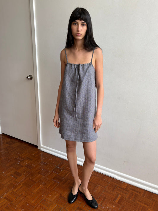 A woman with dark hair and a fringe stands in a room wearing the Weave Mini - Indigo Check by Deiji Studios, an indigo micro-check linen mini dress with thin straps and a drawstring front tie, with black flats.