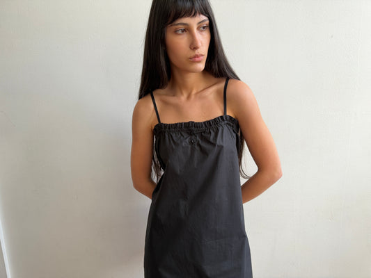 A cropped view of a woman with dark hair and a fringe wearing the Frill Placket Dress - Black by Deiji Studios, a black mini dress with thin straps and a gathered, buttoned frill placket at the neckline.