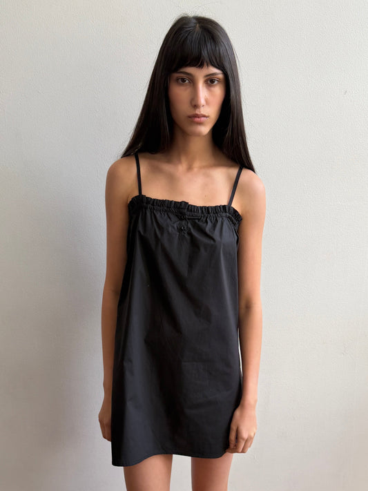 A woman with dark hair and a fringe stands against a wall wearing the Frill Placket Dress - Black by Deiji Studios, a black mini dress with thin straps and a gathered, buttoned frill placket at the neckline.