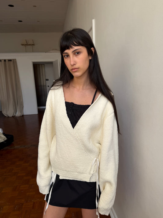 A woman with dark hair and a fringe stands in a room wearing the Frill Placket Dress - Black by Deiji Studios, a black mini dress with a frill placket neckline, layered under a cream wrap cardigan.