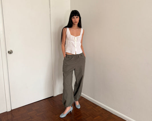 A woman with dark hair and a fringe stands in a room wearing the Relax Tie Top - White by Deiji Studios, a white sleeveless linen top with a drawstring tie neckline, with khaki trousers and pale blue heels.