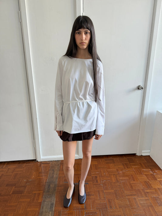 A woman with dark hair and a fringe stands in a room wearing the Wided Longsleeve - White by Deiji Studios, a white top with a wide neckline and drawstring hem, with a brown mini skirt and black flats.