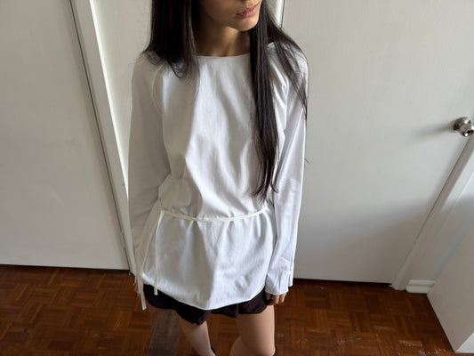 A cropped view of a woman with dark hair and a fringe wearing the Wided Longsleeve - White by Deiji Studios, a white top with a wide neckline and drawstring hem.