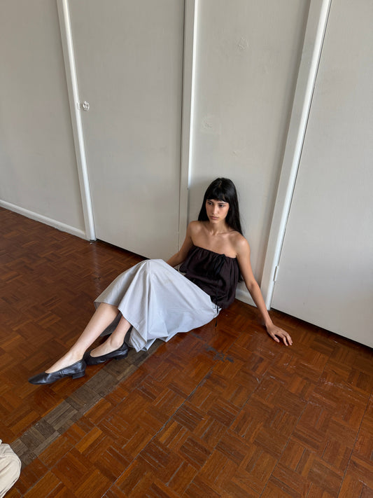 A woman with dark hair and a fringe sits on the floor against a door wearing the Wrap Skirt - Cloud Stripe by Deiji Studios, a pale grey-blue fine-striped wrap midi skirt, with a brown strapless bubble top.
