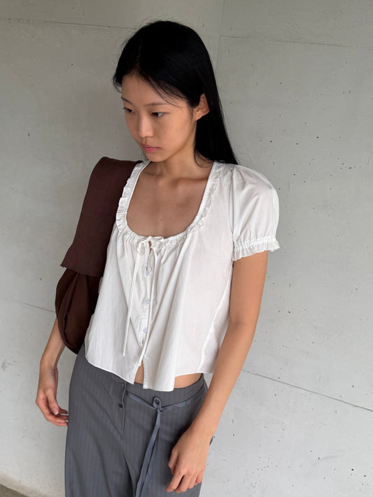 A woman with long dark hair leans against a concrete wall wearing the Flounced Top - White by Deiji Studios, a white top with a frilled scoop neckline, front tie and puff sleeves, with grey pinstripe trousers and a brown shoulder bag.