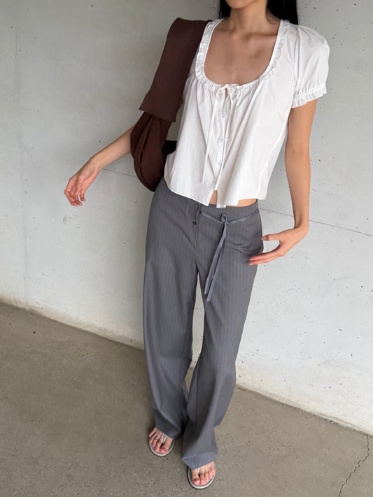A woman with long dark hair stands against a concrete wall wearing the Flounced Top - White by Deiji Studios, a white top with a frilled scoop neckline and puff sleeves, with grey pinstripe trousers and a brown shoulder bag.