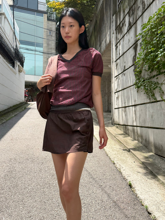 A woman with long dark hair walks along a sunlit lane wearing the Cross Neck Tee - Red Stripe by Deiji Studios, a fine knit tee with red and charcoal vertical stripes and a V-neckline, paired with a brown mini skirt and a shoulder bag.