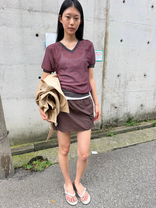 A woman with long dark hair stands against a concrete wall holding a beige jacket, wearing the Cross Neck Tee - Red Stripe by Deiji Studios, a fine red and charcoal striped knit tee, with a brown mini skirt.