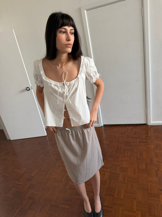 A woman with dark hair and a fringe stands in a room wearing the Flounced Top - White by Deiji Studios, a white top with a frilled scoop neckline, front tie and puff sleeves, with a pale grey skirt and black flats.