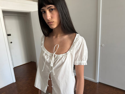 A cropped view of a woman with dark hair and a fringe wearing the Flounced Top - White by Deiji Studios, a white top with a frilled scoop neckline, front tie and puff sleeves, in a room.