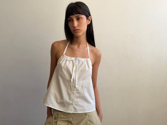 A cropped view of a woman with dark hair and a fringe wearing the Simple Tie Top - White by Deiji Studios, a white halter top with a gathered neckline and front tie, with cream trousers.
