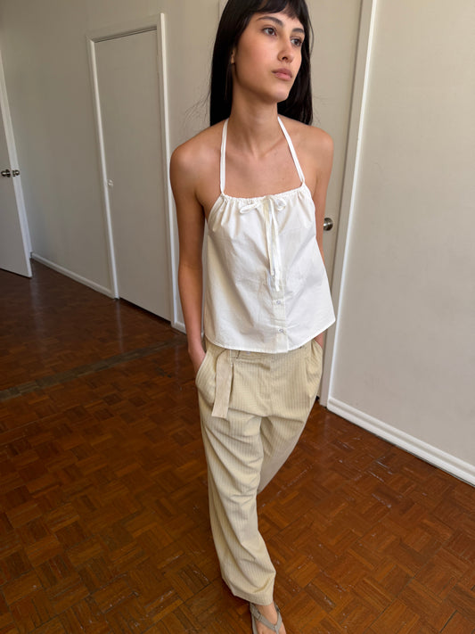A woman with dark hair and a fringe stands in a room wearing the Simple Tie Top - White by Deiji Studios, a white halter top with a gathered neckline and front tie, with cream trousers.