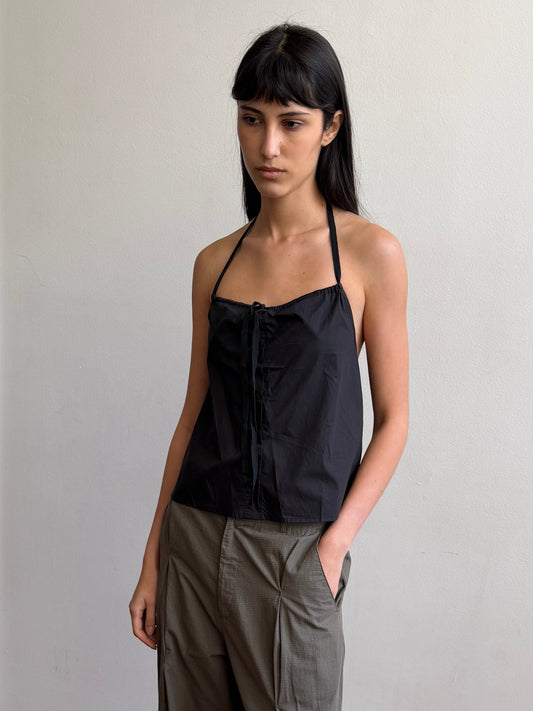 A woman with dark hair and a fringe stands in a room wearing the Simple Tie Top - Black by Deiji Studios, a black halter top with a gathered neckline and drawstring front tie, with khaki trousers.