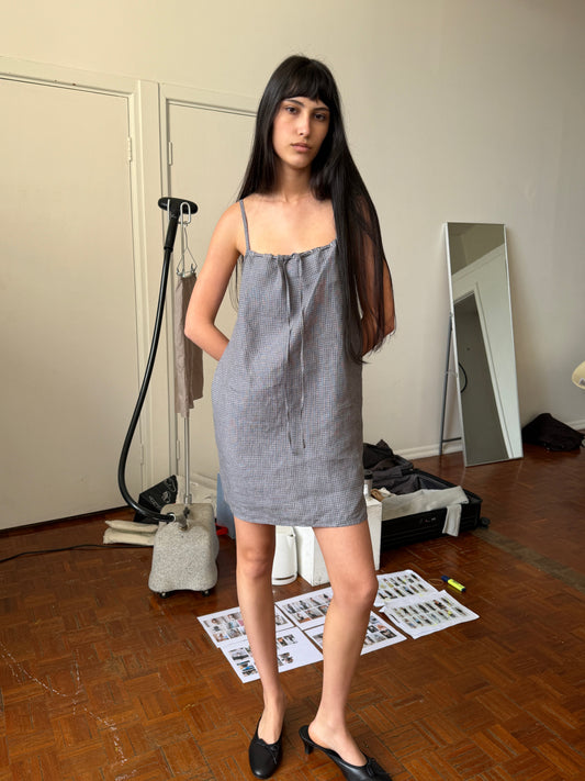 A woman with dark hair and a fringe stands in a room wearing the Weave Mini - Indigo Check by Deiji Studios, an indigo micro-check linen mini dress with thin straps, and black heels.