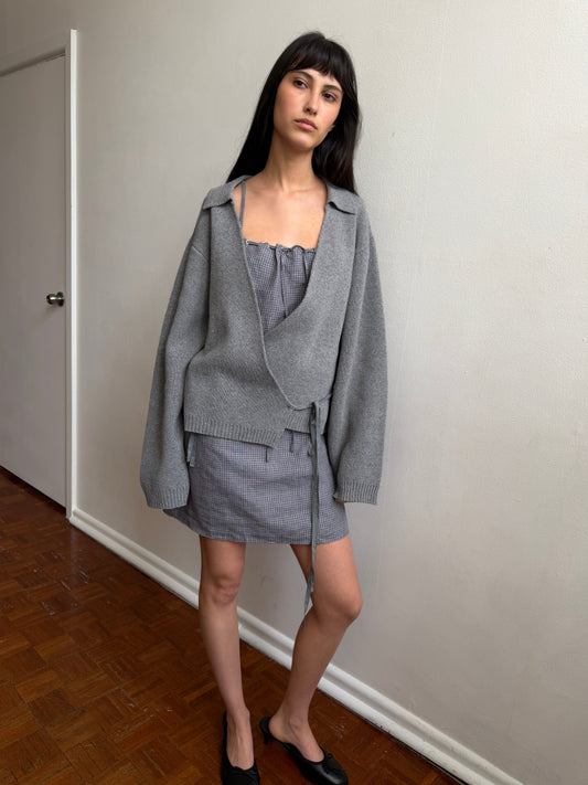 A woman with dark hair and a fringe stands in a room wearing the Wrap Cardigan - Grey by Deiji Studios, a grey knit wrap cardigan with a collar and waist tie, over a grey top and mini skirt with black flats.