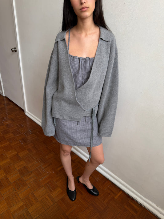 A woman with dark hair and a fringe stands in a room wearing the Weave Mini - Indigo Check by Deiji Studios, an indigo micro-check linen mini dress, layered under a grey wrap cardigan with black flats.