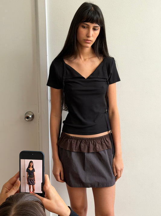 A woman with dark hair and a fringe stands against a door wearing the Flounce Layer Mini Skirt - Brown Blue Stripe by Deiji Studios, a fine brown-blue stripe mini skirt with a brown flounce waist, and a black tee, as another person photographs her on a phone.
