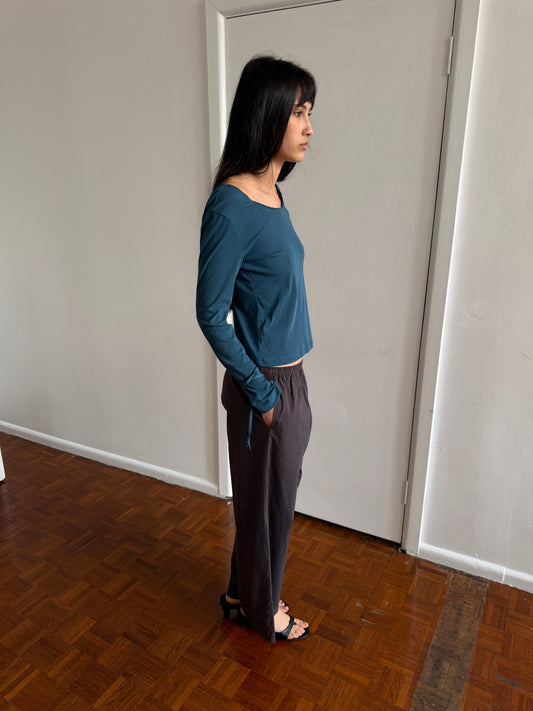A woman with dark hair and a fringe stands in profile in a room wearing the Ease Trouser - Brown Blue Stripe by Deiji Studios, dark brown wide-leg trousers with a fine blue pinstripe, with a teal long-sleeve top and black sandals.