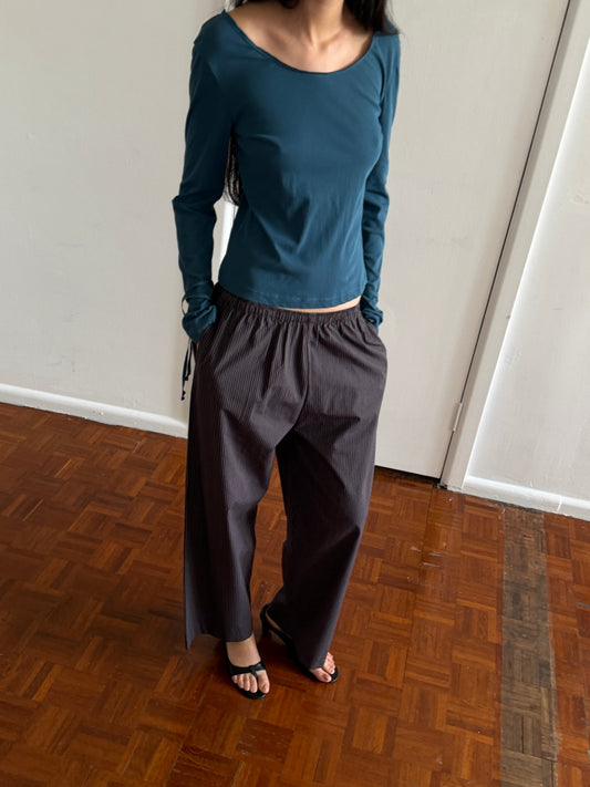 A cropped view of a woman wearing the Ease Trouser - Brown Blue Stripe by Deiji Studios, dark brown wide-leg trousers with a fine blue pinstripe, with a teal long-sleeve top and black sandals, in a room.