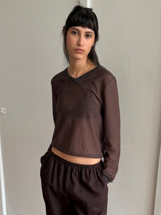 A woman with long dark hair and bangs wears the Stripe Longsleeve in Brown Grey Stripe, paired with matching pants, standing against a plain light-colored wall with her hands in her pockets.