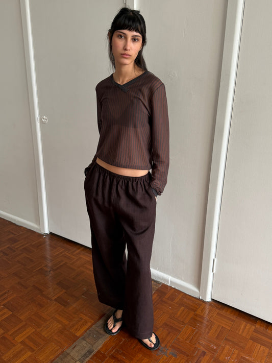 A woman with dark hair and a fringe stands in a room with hands in her pockets, wearing the Linen Ease Trouser - Brown Black Stripe by Deiji Studios, dark brown fine-striped wide-leg linen trousers, with a matching brown striped knit top and black flip-flops.