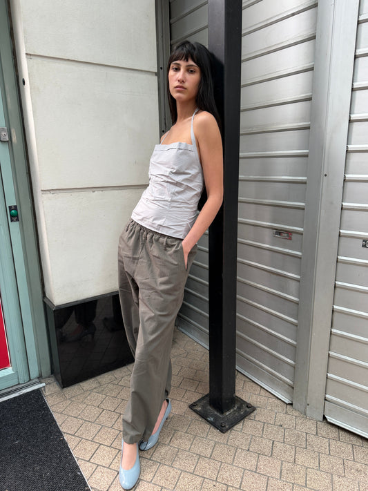 A woman with dark hair and a fringe leans against a post on a street wearing the Folded Top - Cloud Stripe by Deiji Studios, a pale grey-blue fine-striped fold-over halter top, with grey trousers and pale blue heels.