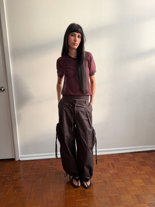 A woman with dark hair and a fringe stands against a wall in a room, wearing the Cross Neck Tee - Red Stripe by Deiji Studios, a fine red and charcoal striped knit tee, with dark brown wide-leg trousers and black sandals.