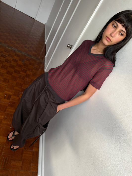 A woman with dark hair and a fringe leans against a wall in a room, wearing the Cross Neck Tee - Red Stripe by Deiji Studios, a fine red and charcoal striped knit tee, with dark brown wide-leg trousers.