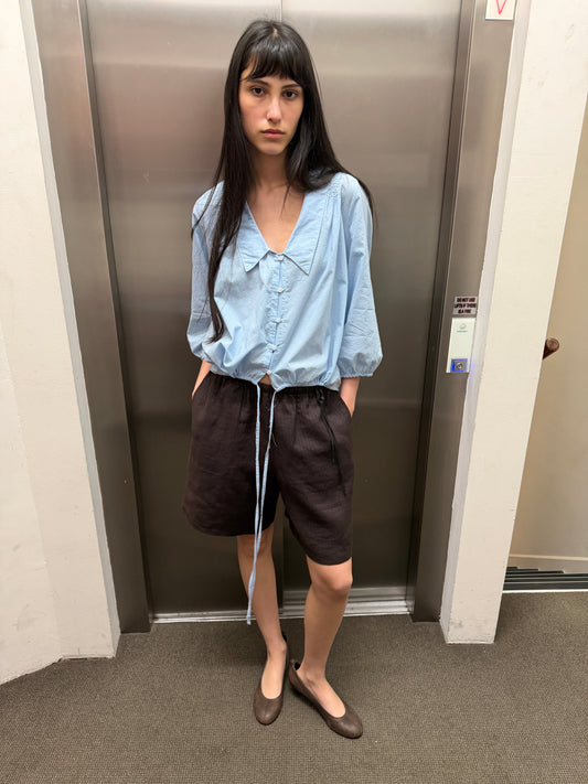 A woman with dark hair and a fringe stands in a lift wearing the Half Short - Brown Black Stripe by Deiji Studios, knee-length dark brown striped linen shorts, with a pale blue collared top and brown flats.