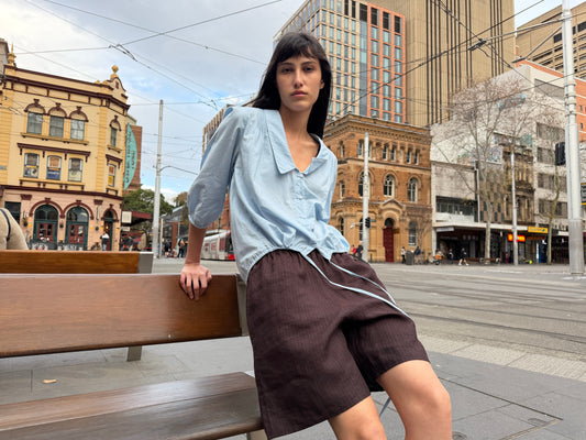 A woman with dark hair and a fringe sits on a bench in a city square wearing the Half Short - Brown Black Stripe by Deiji Studios, knee-length dark brown striped linen shorts, with a pale blue collared top.