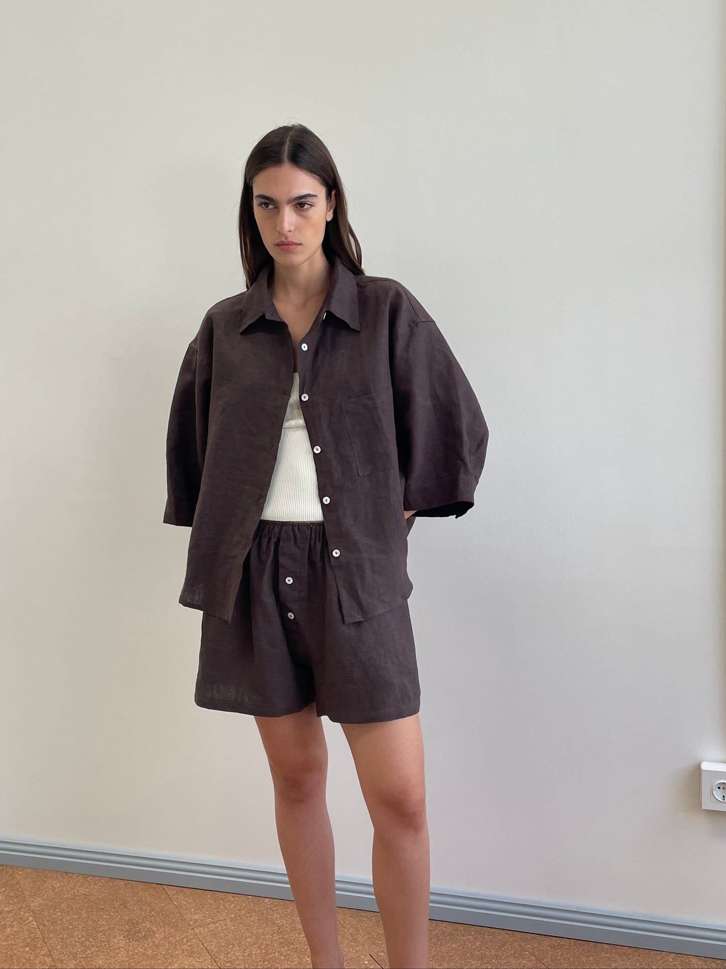 A woman with straight dark hair and a neutral expression stands indoors against a beige wall, wearing the 03 Set - Bark: a loose organic linen dark brown shirt over a white top and matching boxer-style shorts.
