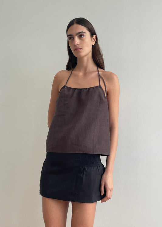 A woman with long brown hair wears a loose dark brown halter top and the Shirring Skirt, standing against a plain light-colored wall. She looks slightly to the side with a neutral expression.