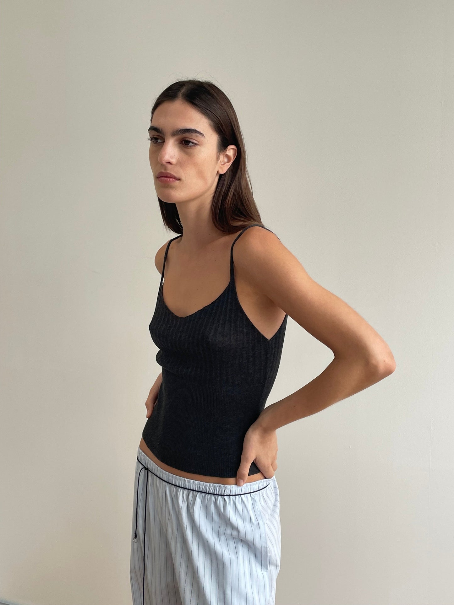 A woman with long brown hair wears the Deiji Studios Ribbed Tank in Dark Charcoal and light blue striped lyocell pants, standing against a plain light wall and looking to the side with a neutral expression.