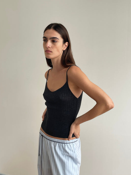 A woman with long brown hair wears the Deiji Studios Ribbed Tank in Dark Charcoal and light blue striped lyocell pants, standing against a plain light wall and looking to the side with a neutral expression.