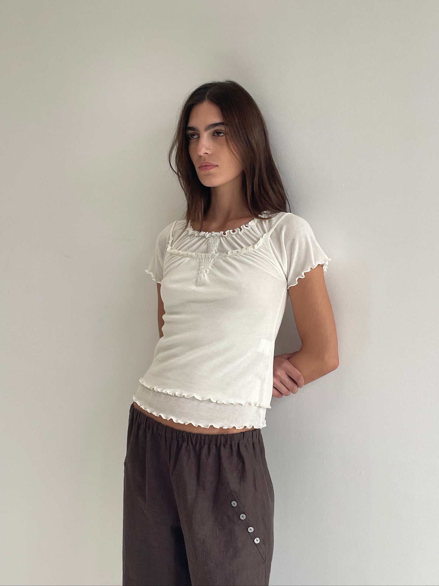 A woman with long brown hair leans against a white wall, wearing a fitted white ruffled-edge top and the Angle Pocket Pant - Bark, which are relaxed, mid-waist pants made from organic linen. She gazes to the side with a neutral expression.