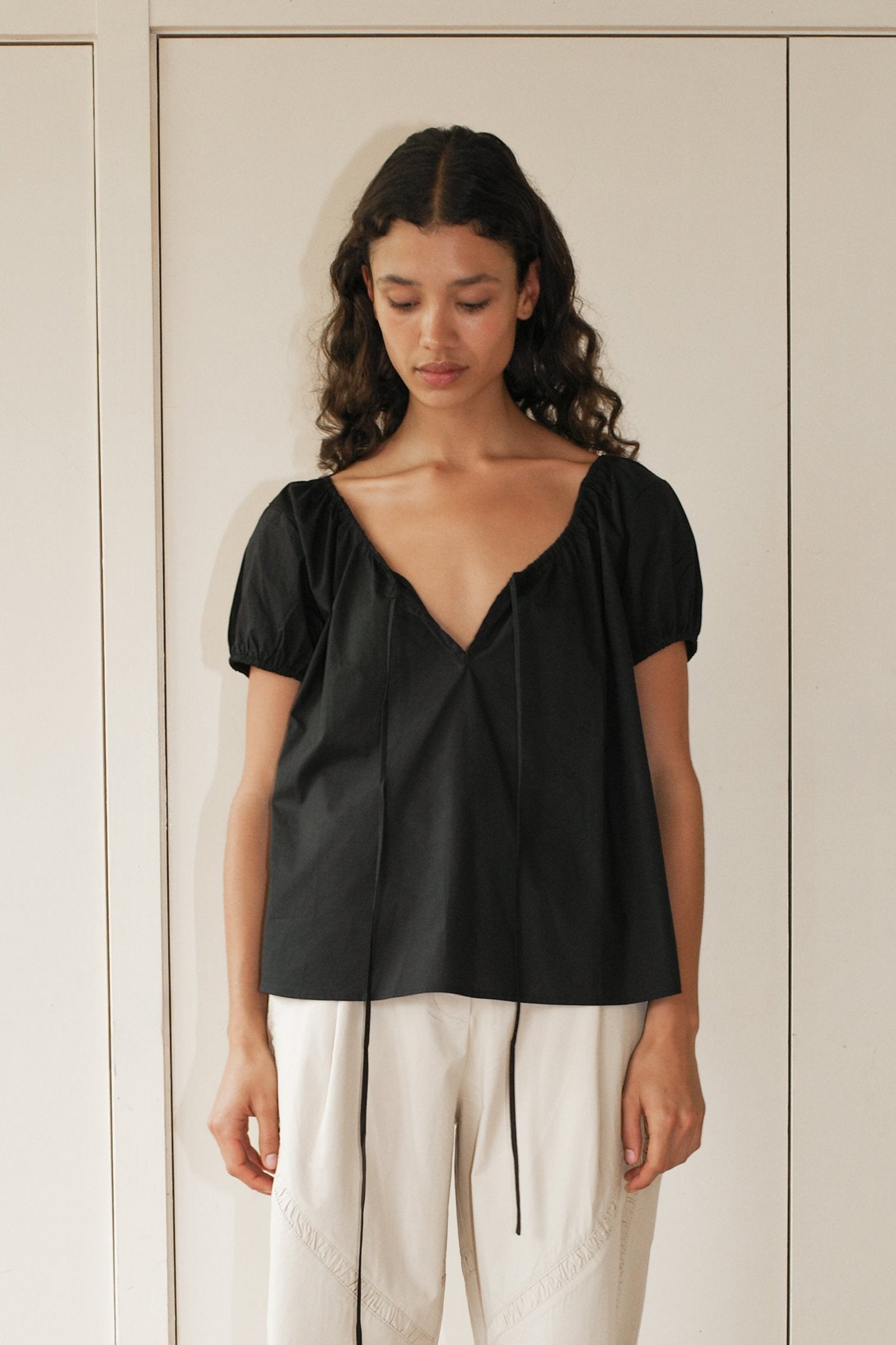 A woman with dark curly hair wears the One Panel Top, a loose black short-sleeve V-neck blouse, paired with white high-waisted pants. She stands indoors against a pale wall, looking down with a neutral expression.