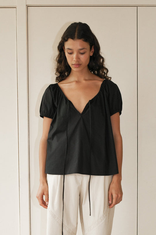 A woman with dark curly hair wears the One Panel Top, a loose black short-sleeve V-neck blouse, paired with white high-waisted pants. She stands indoors against a pale wall, looking down with a neutral expression.