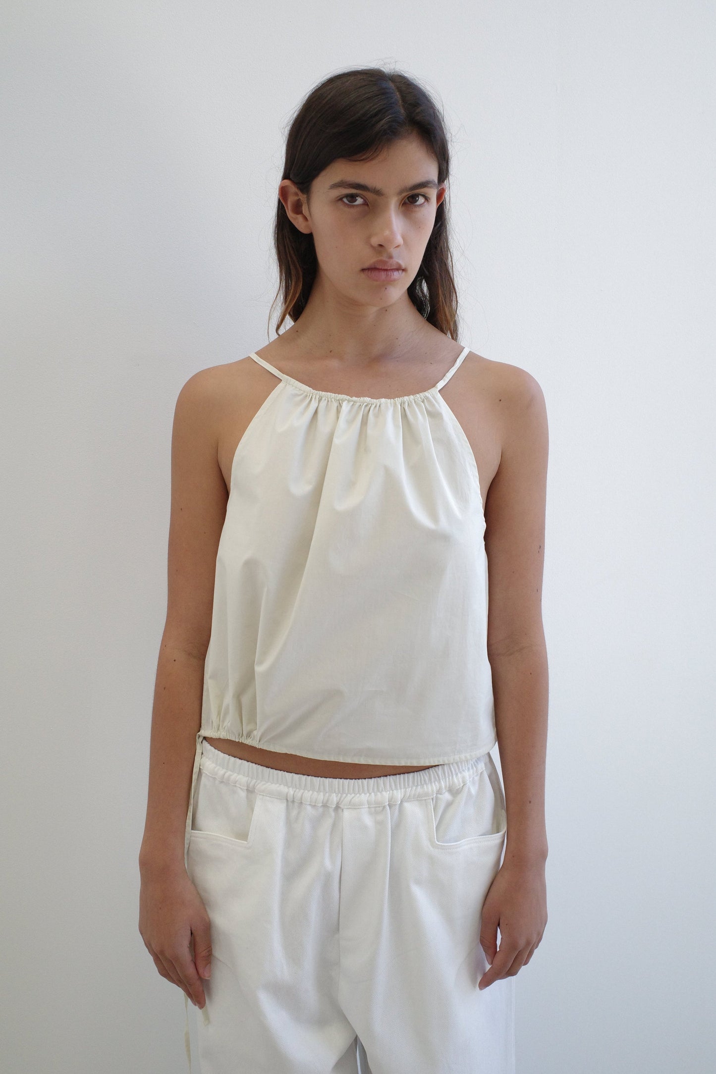 A person with long brown hair wears the Simple Top - Butter White, a sleeveless, loose-fitting organic cotton top by Deiji Studios, paired with white pants, standing against a plain white background with a neutral expression.