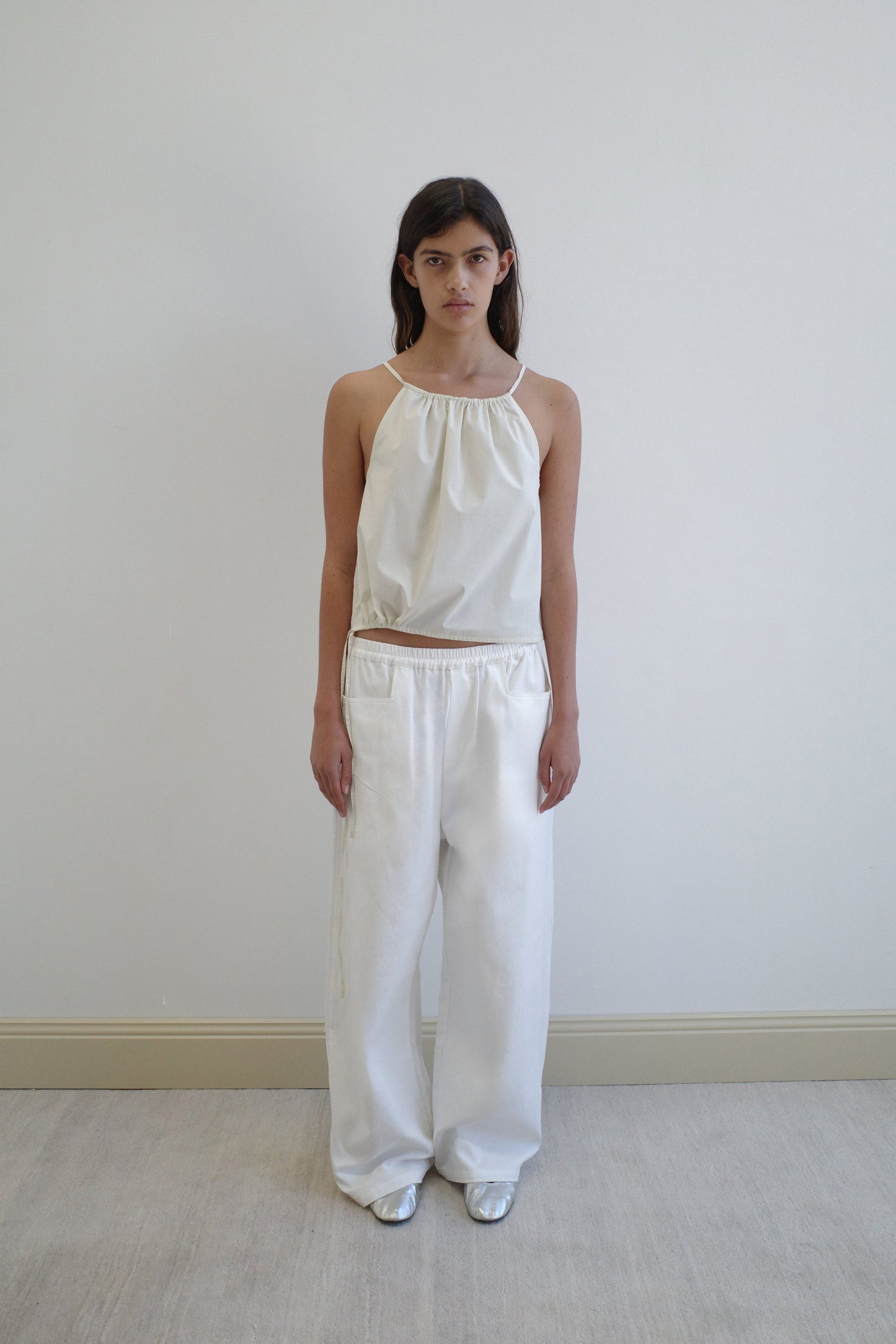 A woman with long dark hair stands against a white wall, wearing the Simple Top - Butter White from Deiji Studios, featuring a loose fit and gathered neckline, paired with wide-legged white pants and silver shoes. She has a neutral expression and relaxed posture.