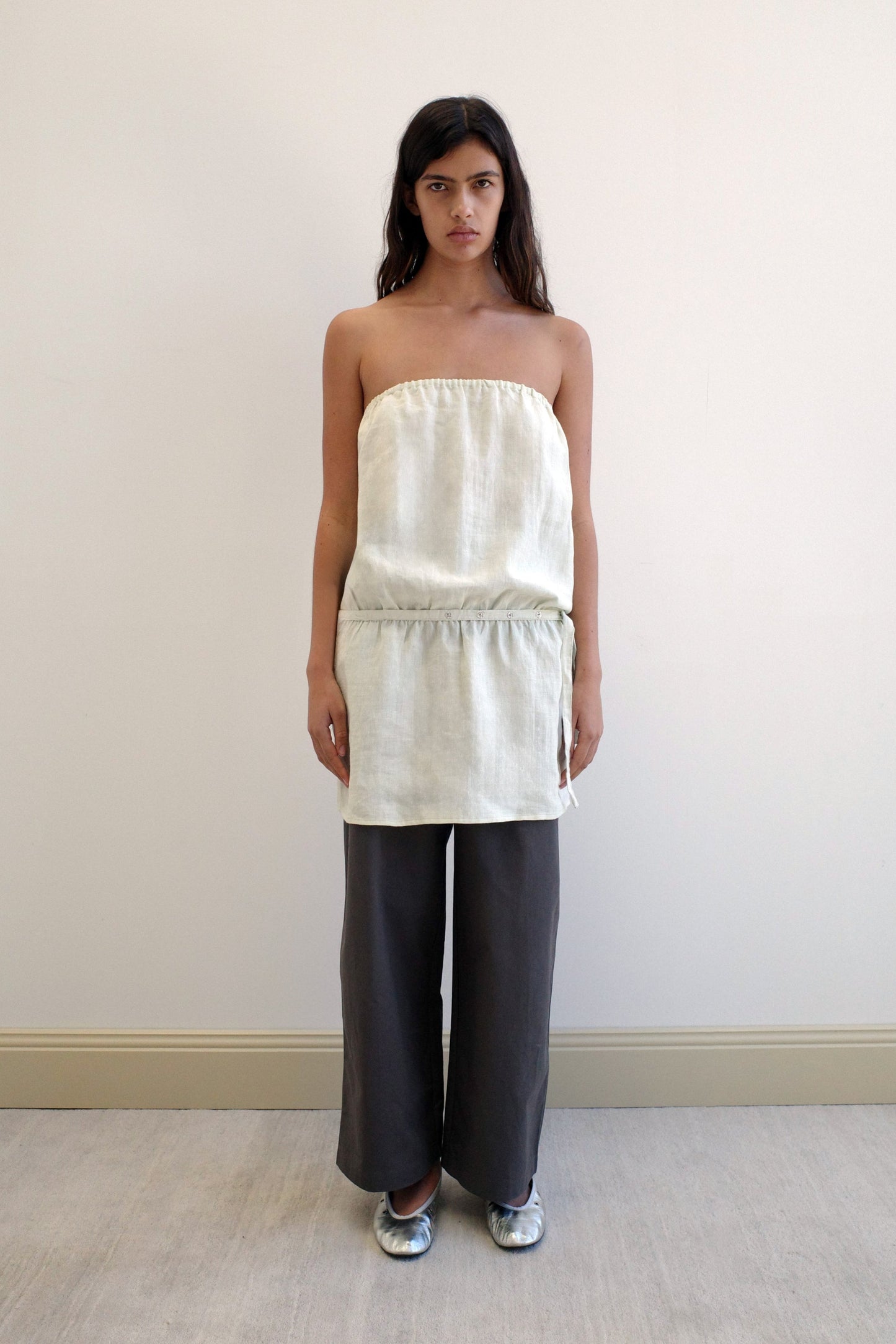 A woman with long dark hair and a neutral expression stands against a beige wall, wearing the Strapless Linen Dress in Pastel Pistachio over wide-leg dark gray pants and silver shoes.