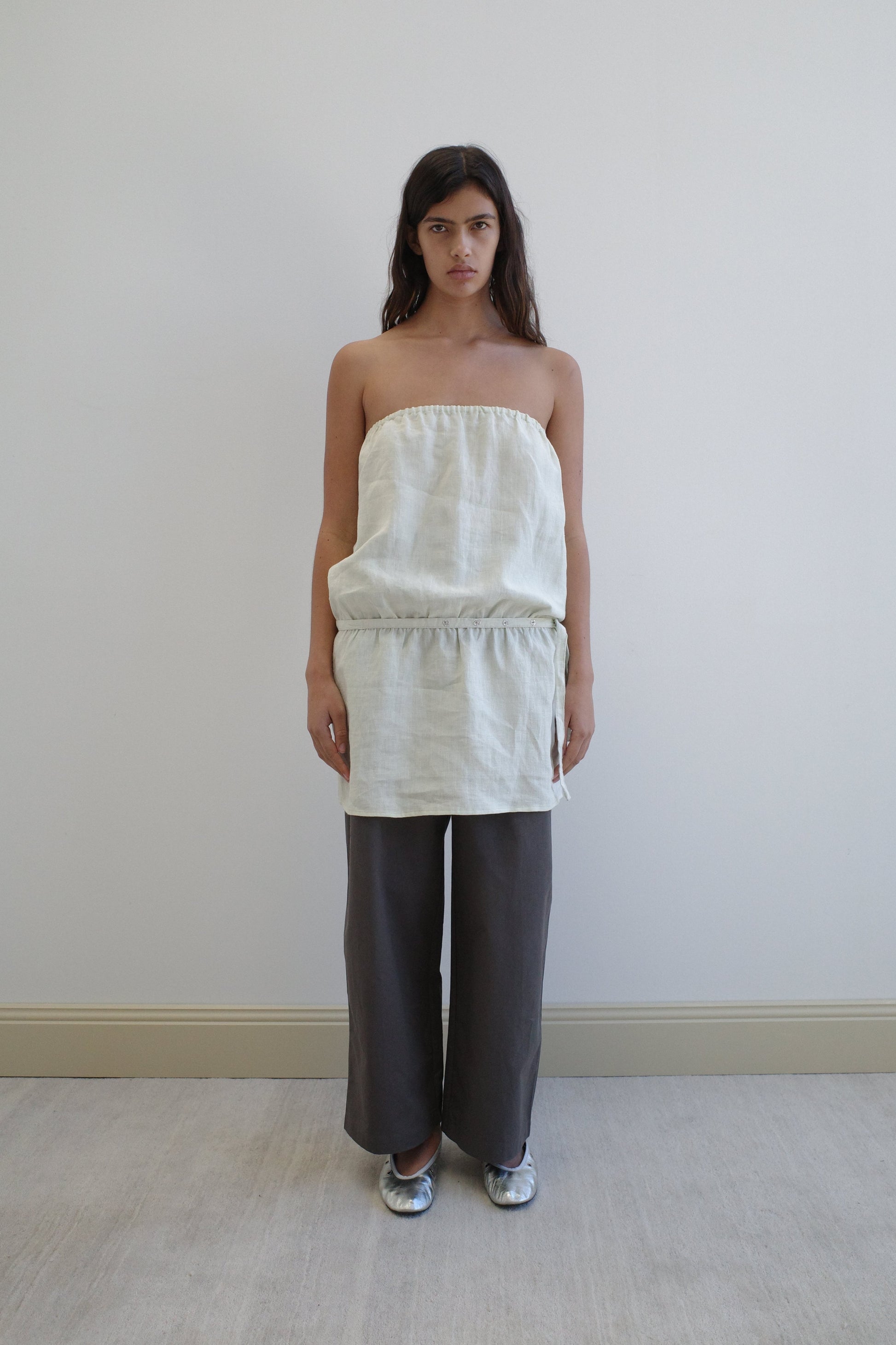 A woman stands against a plain white wall wearing a strapless, light top with a drawstring waist, Deiji Studios Two Layer Pant - Charcoal in a wide-leg silhouette, and silver flat shoes.