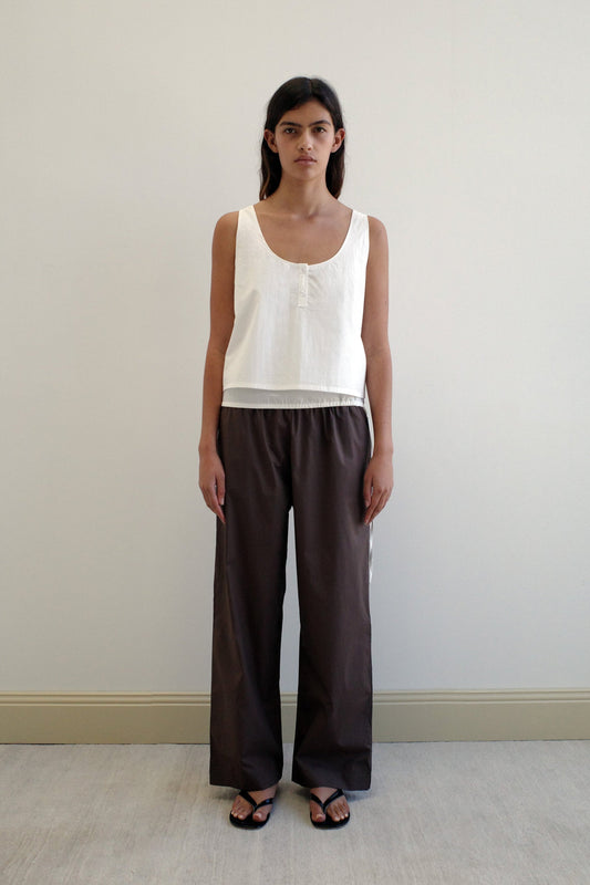 A person stands indoors against a plain light-colored wall, wearing a sleeveless white top and the Ease Trouser - Chocolate by Deiji Studios in organic cotton, paired with black sandals, looking straight at the camera with a neutral expression.