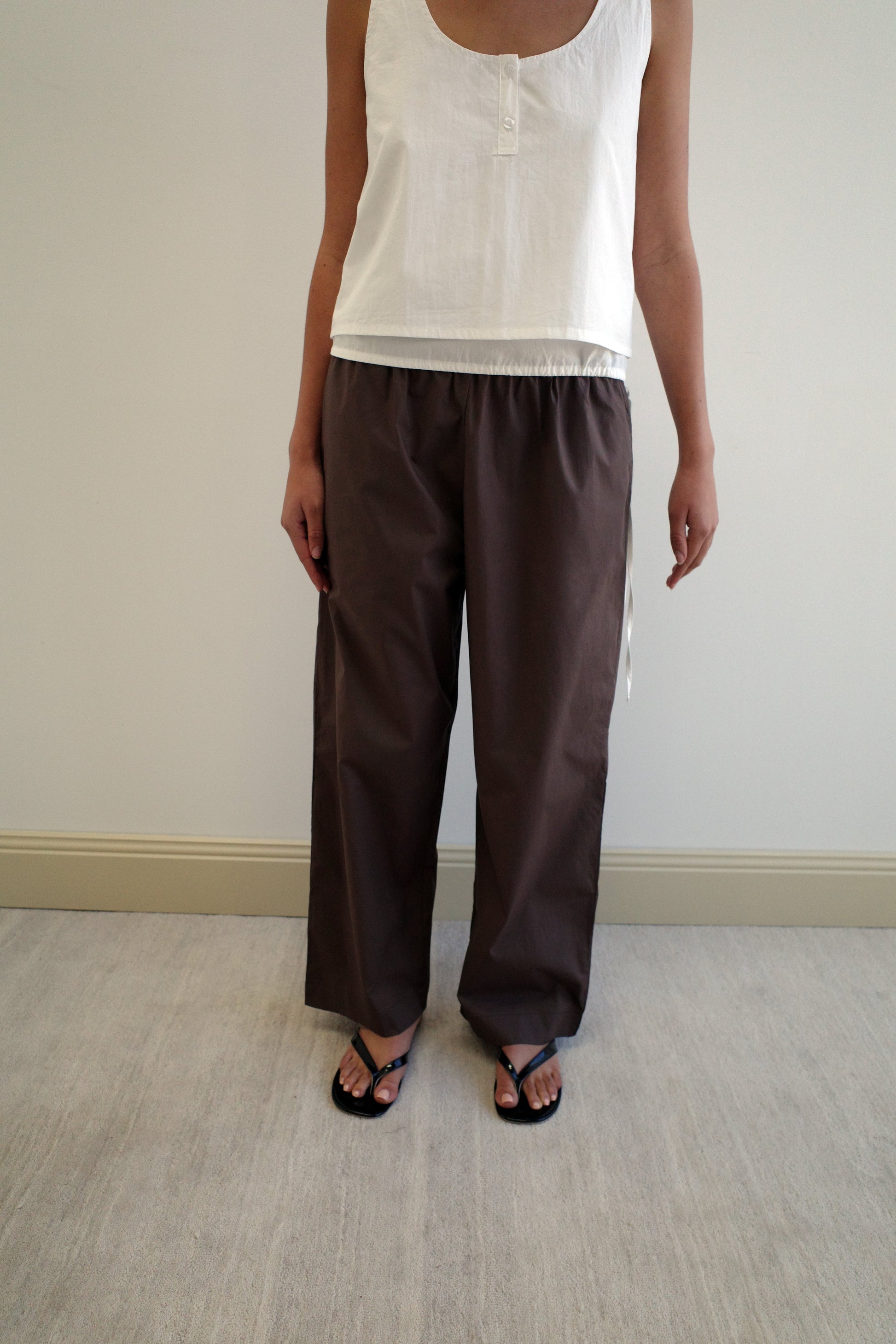 A person stands indoors wearing a sleeveless white top, Deiji Studios Ease Trouser in Chocolate, and black sandals. The cropped photo highlights their torso, legs, and feet against a plain background.