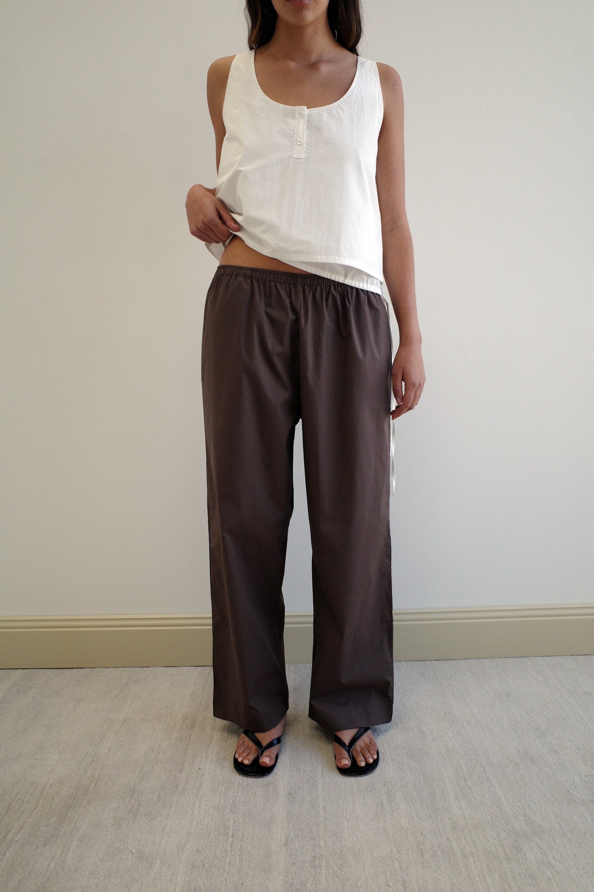 A woman stands indoors against a plain light wall, wearing a sleeveless white top, Deiji Studios Ease Trouser - Chocolate in loose brown organic cotton, and black sandals, with one hand resting on her waist.