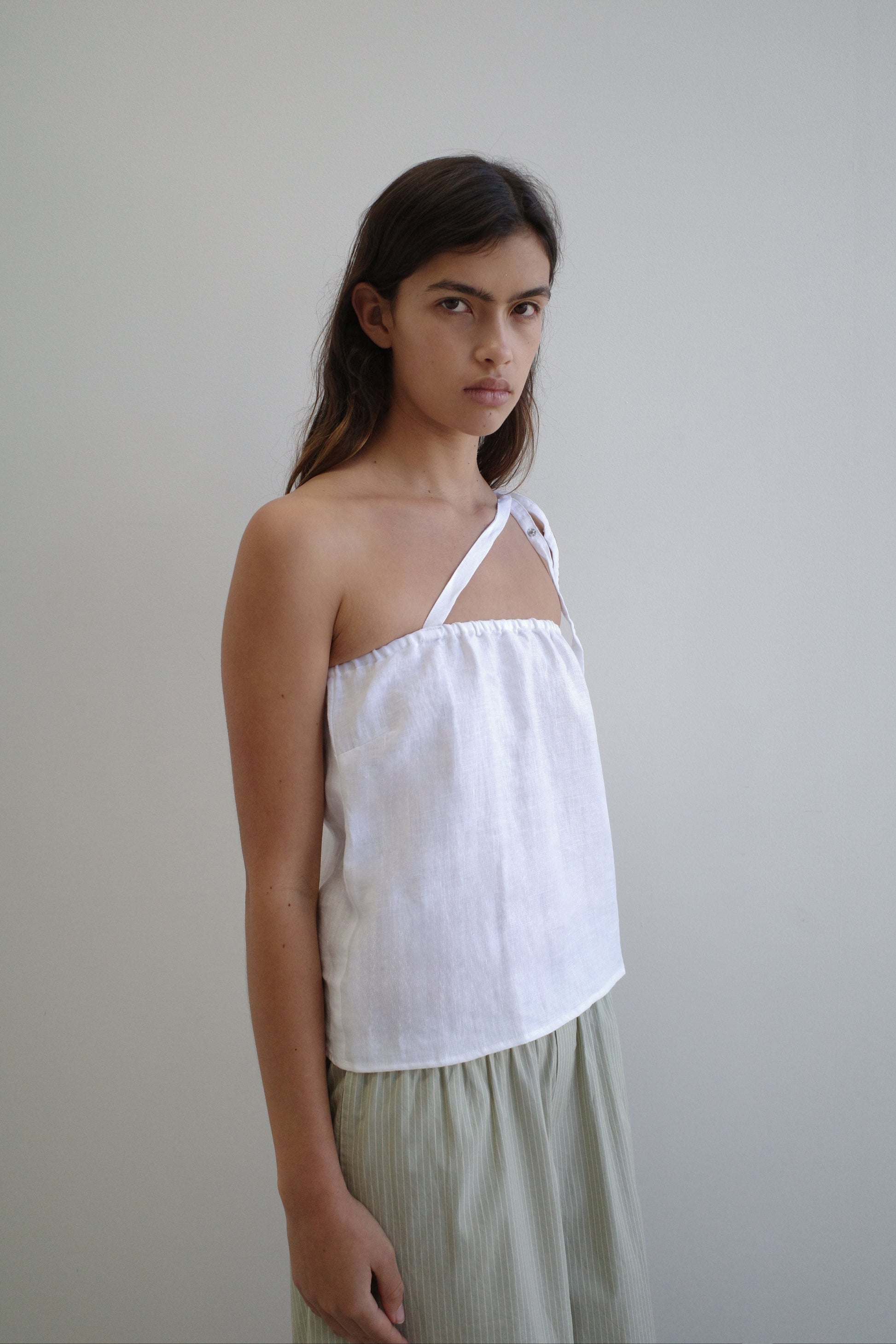 A woman with long brown hair wears the Button Strap Top - White by Deiji Studios and a light pleated skirt, standing against a plain, light-colored background.