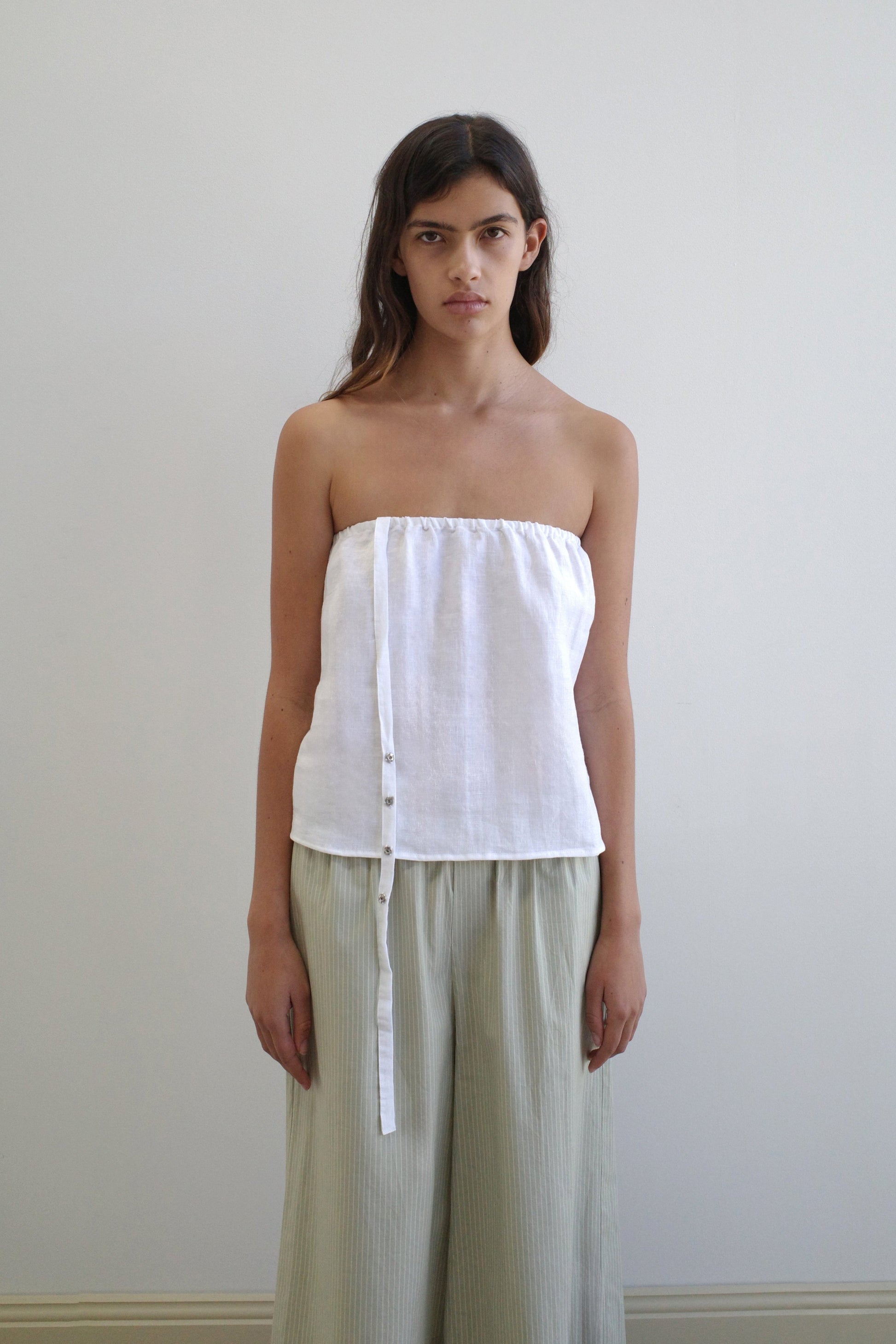 A woman with long brown hair stands against a plain light wall, wearing the Button Strap Top in white from Deiji Studios and loose, light green pants. She faces the camera with a neutral expression.