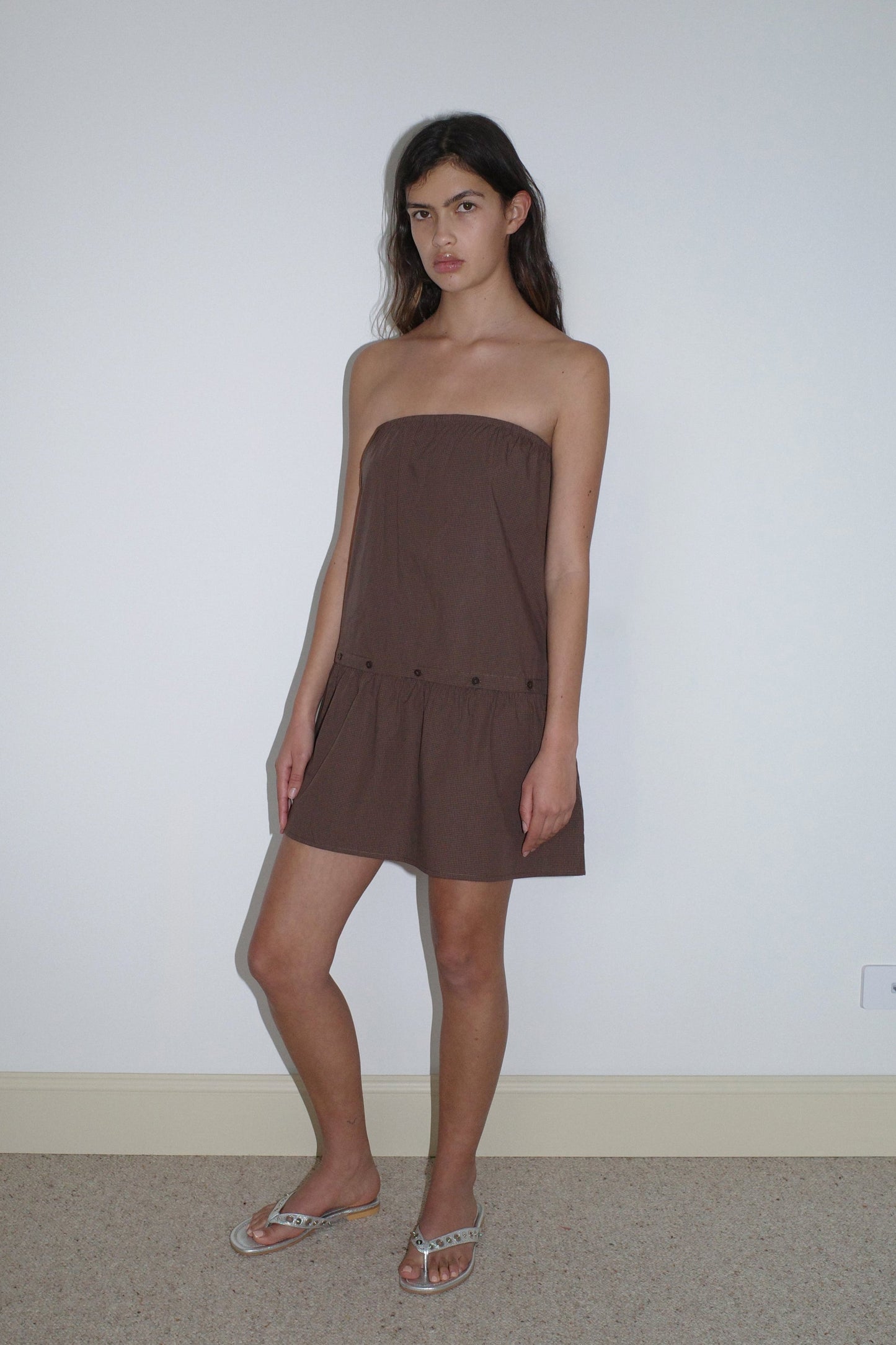 A woman stands indoors against a white wall, wearing the Button Dress - Carob Check from Deiji Studios and silver sandals. She has long dark hair, a neutral expression, and stands with one leg slightly bent.