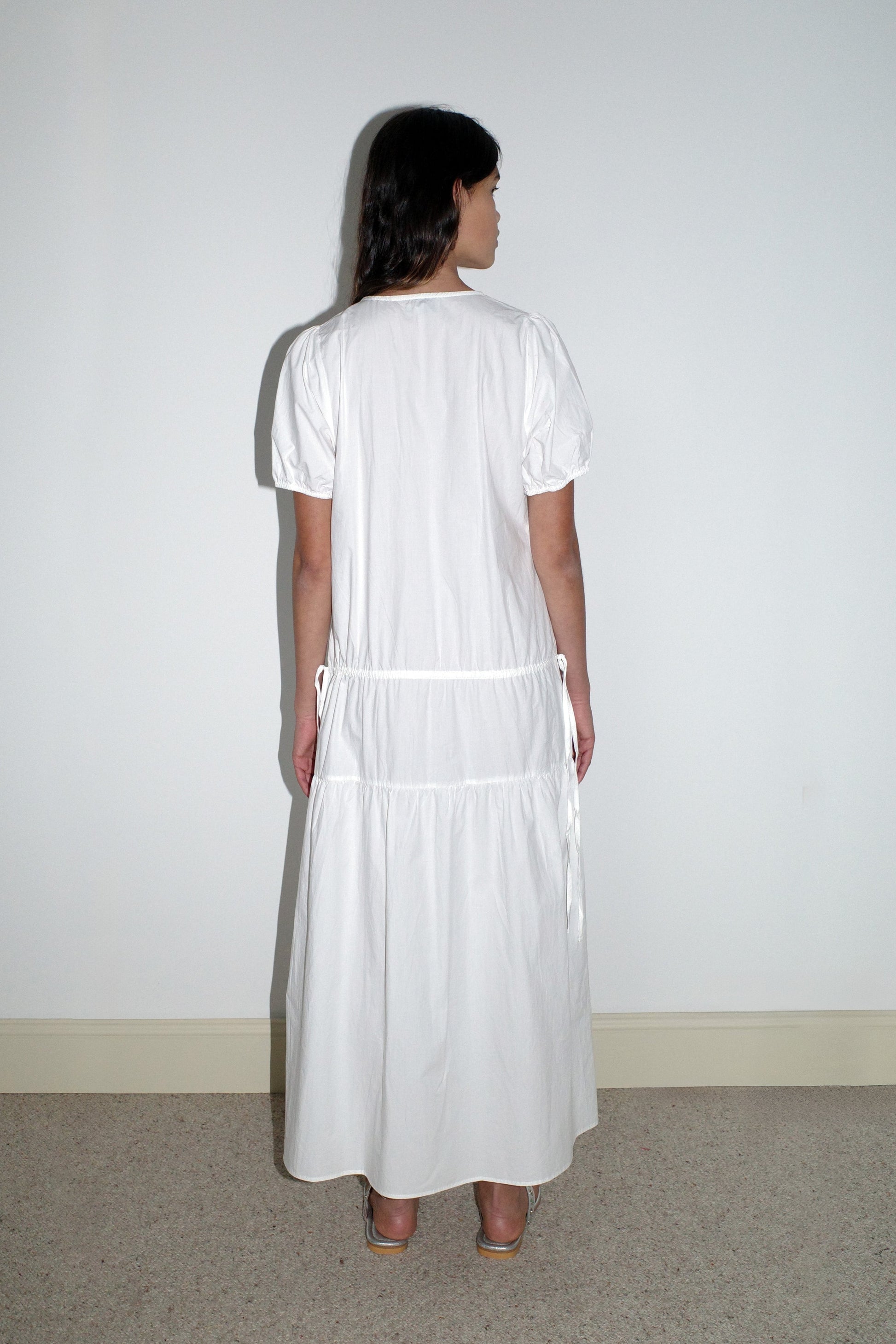A woman stands with her back to the camera on a beige carpet, wearing the Waistcord Dress - White by Deiji Studios—an ankle-length, short-sleeved organic cotton dress—and beige heels against a white wall.