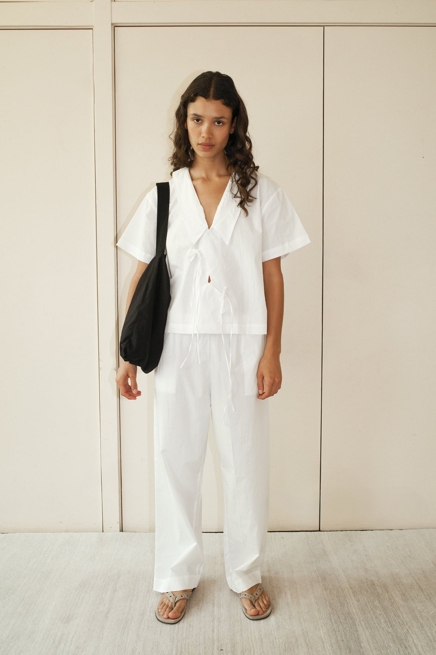 A person with long curly hair wears the Panel Shirt - White from Deiji Studios and matching wide-leg pants, carrying a black shoulder bag and wearing sandals, standing against a light beige wall.