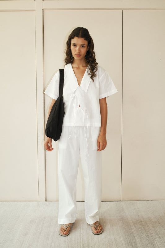 A person with long curly hair wears the Panel Shirt - White from Deiji Studios and matching wide-leg pants, carrying a black shoulder bag and wearing sandals, standing against a light beige wall.